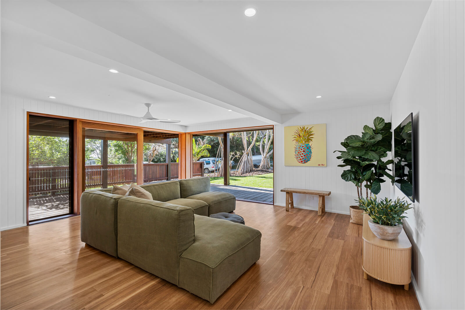 Bright coastal living room with timber floors, green sofa, indoor plants, and large windows opening to a leafy outdoor deck.