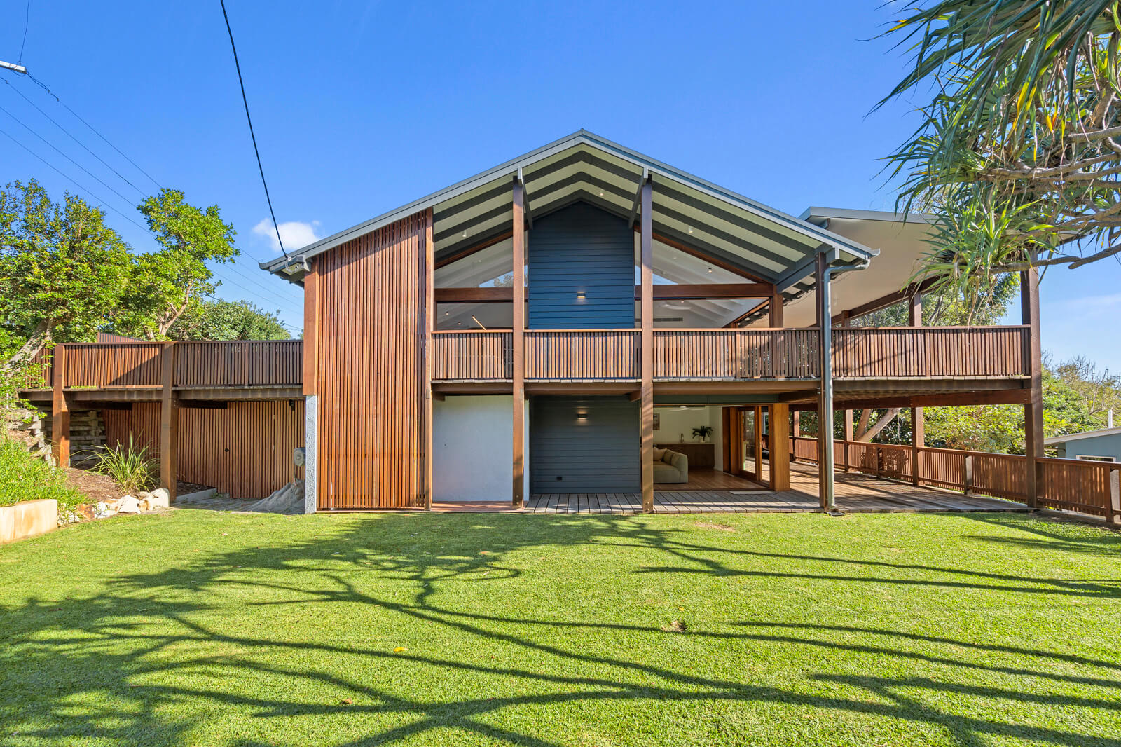 Modern coastal holiday home on Stradbroke Island with timber deck, lawn, gabled roof, and lush tropical surroundings.
