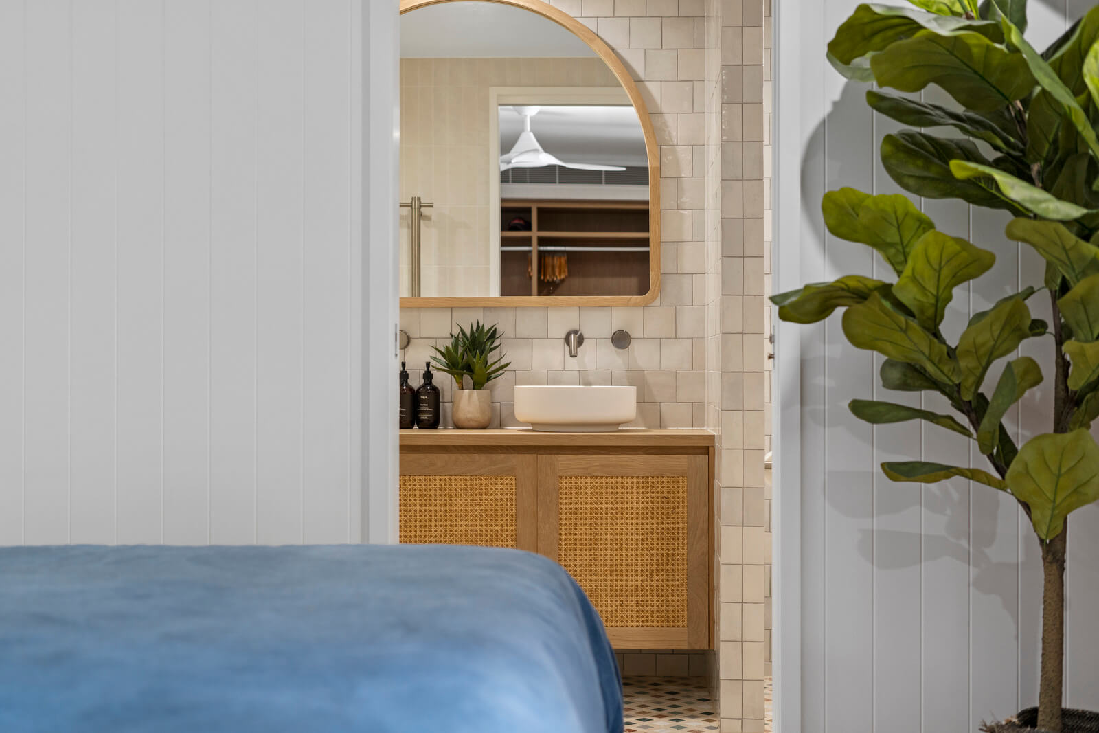 Coastal bedroom view featuring timber vanity, curved mirror, tiled bathroom, soft blue bedding, and indoor greenery.