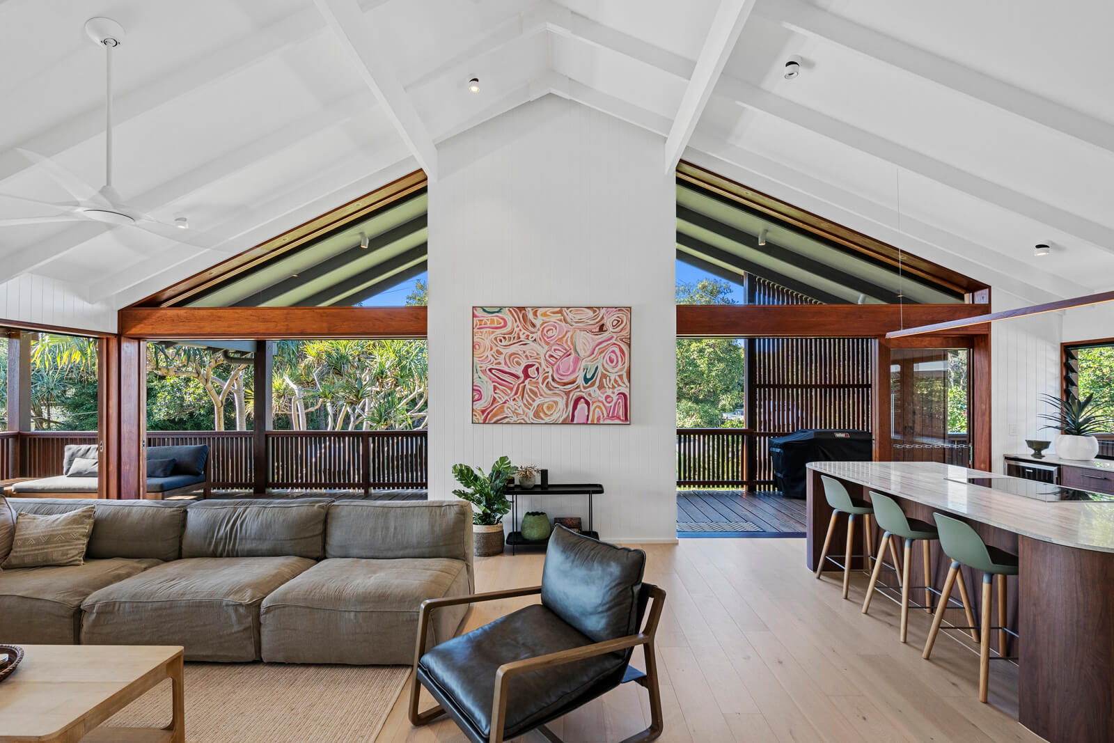 Modern coastal home interior with vaulted white ceiling, timber beams, open kitchen, lounge area, and leafy outdoor views.